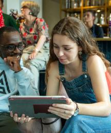A young man sitting next to a young woman who is holding a tablet which they are both looking at