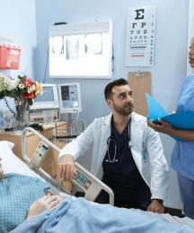 A female nurse standing beside a hospital female patient's bed, holding a file and talking to the patient. A male doctor is sitting beside the bed listening to the nurse talk