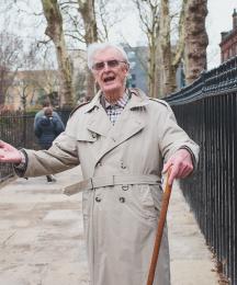 A man in a rain mac holding a walking stick holding his arms out