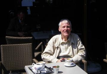 An elderly man sitting at a cafe table, looking at the camera and smiling