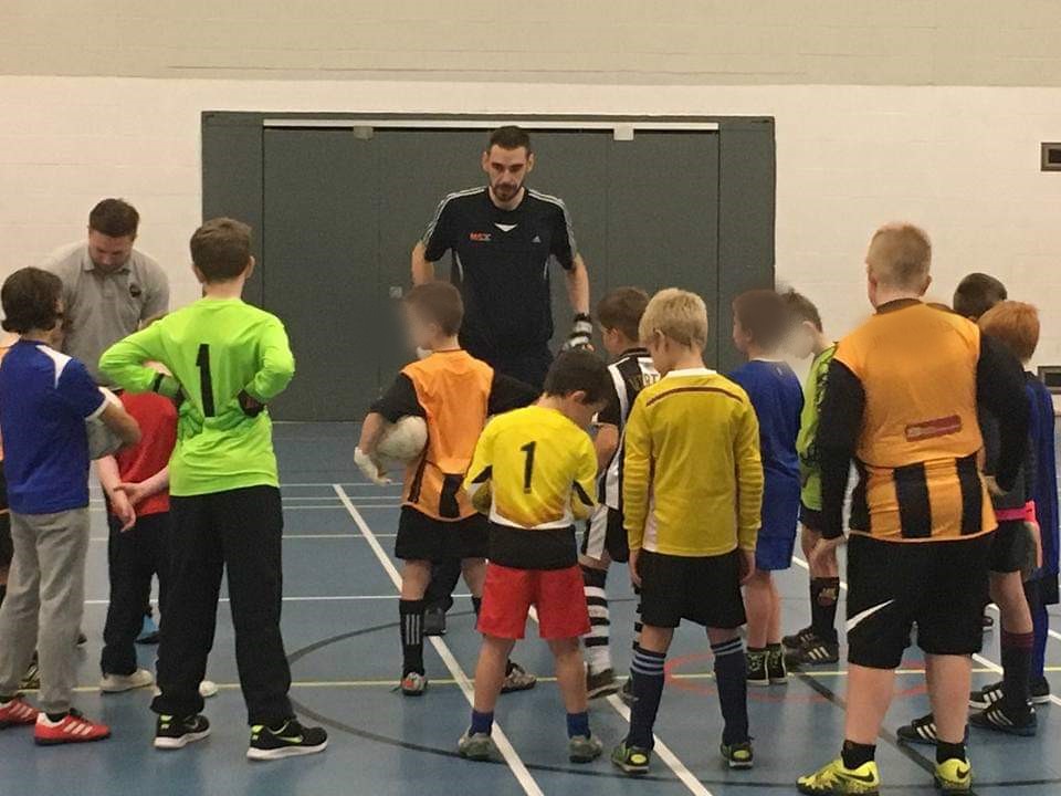 A man in a sports hall with a group of young children in football gear