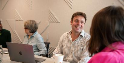 A man sitting at a desk chatting to a woman opposite whose face we can't see, with another woman sitting next to him chatting with a man next to her