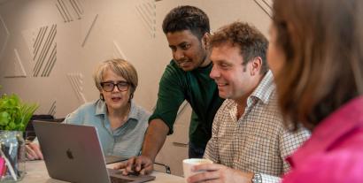 A man standing and demonstrating something on a laptop, watched by a man and a woman sitting either side.