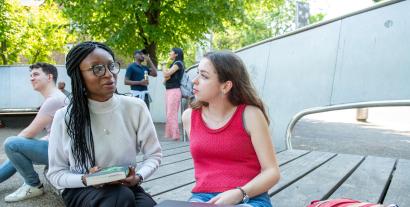 Two women seated in an outside setting and chatting, with other people in the background