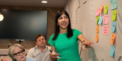 A woman standing beside a wall presenting to two people sitting beside her