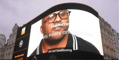 An advertising screen in Piccadilly Circus displaying a man looking at the camera, with the words 'Stamma' on it