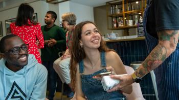 A young man and woman in a cafe looking at a waitress and holding a mobile phone to a card reader