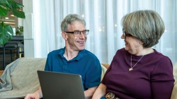 A man and a woman sitting on a sofa laughing, the woman with a laptop on her lap