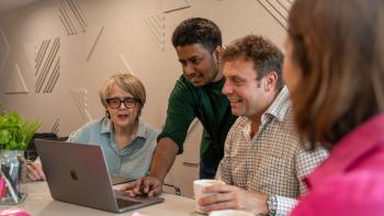 A man standing and demonstrating something on a laptop, watched by a man and a woman sitting either side.