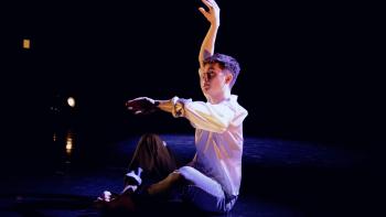 A man sitting on a stage, gesticulating with his arms, mid-dance