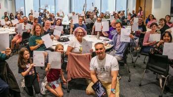 A group of people of all ages in a classroom, smiling and holding paper in the air