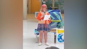 A woman standing with a microphone in her hand, looking at a sheet of paper she's holding