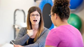 A woman in a therapy setting, pointing at her throat and opening her mouth. Opposite her is a younger woman