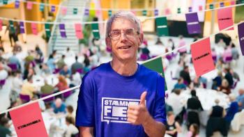 A man smiling and raising his thumb, aganist a backdrop of bunting and a crowded room