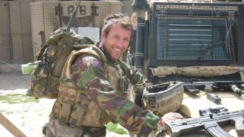 A man in a soldier's uniform standing over a car and smiling