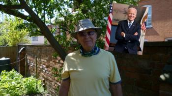 A man wearing a hat and sunglasses, standing in a garden, beside an image of President Joe Biden