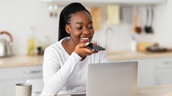 A woman sitting at a kitchen table in front of a laptop and speaking into a smartphone in her hand