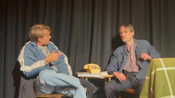 Two male actors, one younger and one older, sitting on a stage and talking in character.