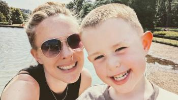 A woman wearing sunglasses standing next to her young son, in front of a lake. Both are smiling