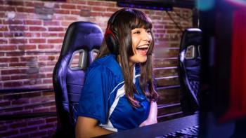 A woman at a desk wearing headphones, looking at a computer screen and laughing