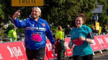 A man and woman running in a sponsored run. The man is holding a phone and taking a selfie