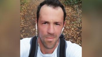 A man in a woodland setting, with earphones in and smiling