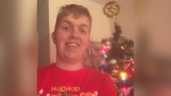 A man wearing a Christmas jumper, standing in front of a Christmas tree, smiling