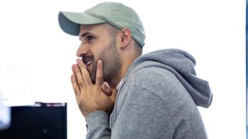 A man in profile, wearing a baseball cap and stroking his chin with his hands