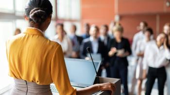 A woman standing a podium and speaking to an audience of people who are standing up