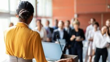 A woman standing a podium and speaking to an audience of people who are standing up