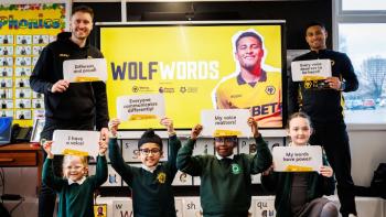 Two men standing either side of a screen, with four schoolchildren kneeling, all holding up paper with writing on it