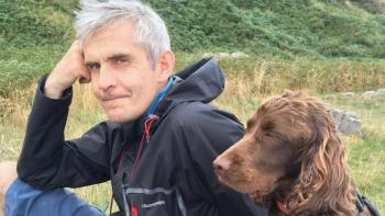 A man sitting on a hillside, resting his head on his hand, with his dog sitting next to him