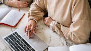 A typing on a laptop whilst holding a pen. Beside her is an other person. We can't see their faces.