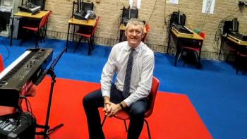 A man sitting in a classroom and smiling, next to a music keyboard and in front of a row of desks
