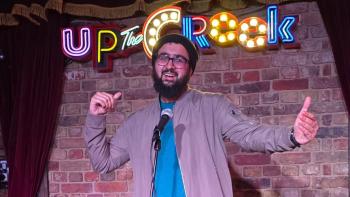 A man on a stage, smiling with his arms outstretched, in front of a microphone. The words 'Up the crook' are in lights behind him