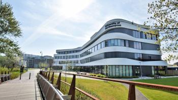 A modern university building surrounded trees and grass, with a bridge leading to it