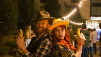 A man and a woman in cowboy outfits standing with their backs to each other and holding up bananas instead of guns