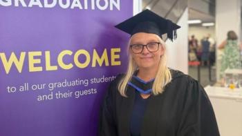 A woman in university gown and mortar board standing in front of a sign saying 'Welcome'