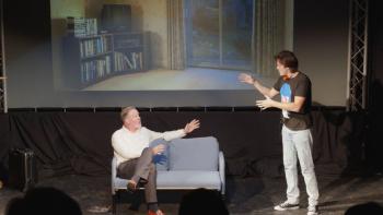 Two male actors on a stage, one sitting on a sofa and the other standing