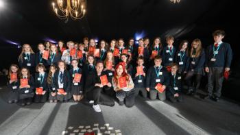 A group of schoolchildren along with a man and a woman posing for a photo, all holding up a brochure