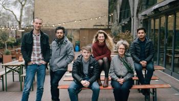 A group of people standing beside and sitting on a bench outdoors and looking at the camera