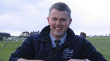 A man standing beside a hedge on a racecourse, smiling