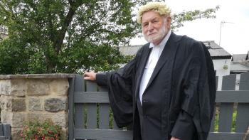 A man in a barrister's outfit leaning on a fence and smiling