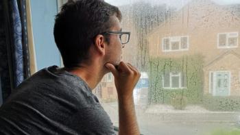 A young man looking out of a window on a rainy day, with his hand to his chin