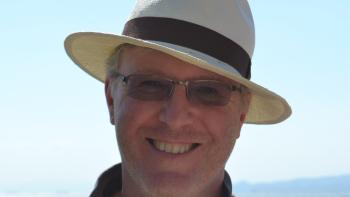 A man in a hat standing with the sea in the background, smiling for the camera