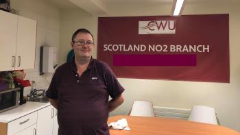 A man in a kitchen in front of a sign saying "CWU - the communications union" and "Scotland No.2 branch"
