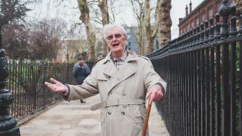 A man in a rain mac holding a walking stick holding his arms out