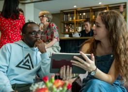 A young man sitting next to and chatting with a young woman who is holding a tablet in a coffee shop