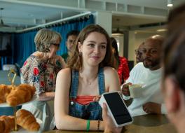 A young woman at a counter in a shop and smiling while using her phone to pay via a card reader held by somebody off screen.
