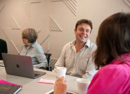 A man sitting at a desk chatting to a woman opposite whose face we can't see, with another woman sitting next to him chatting with a man next to her